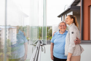 Lasitetulla parvekkeella iäkkäämpi nainen ja nuorempi nainen halaavat toisiaan hymyillen.