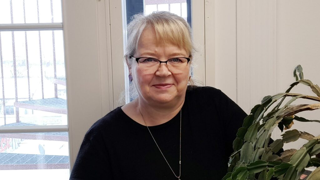 Hymyilevä vaaleahiuksinen silmälaseissa oleva vanhempi nainen päällään musta neule
