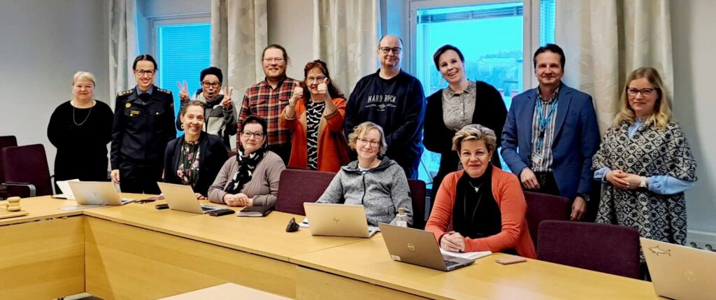 Kuvassa johtoryhmä ensimmäisessä kokouksessaan, pitkän pöydän takana seisomassa ja istumassa miehiä ja naisia.