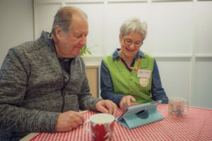 Vapaaehtoistyö Olkan avustaja avustaa ikäihmistä tabletin käytön kanssa pöydän ääressä.