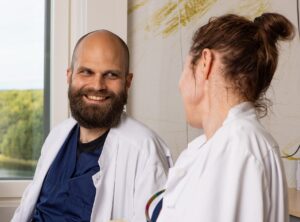 Kaksi valkotakkista lääkäriä hymyilee toisilleen, tummapartainen mies ja tummahiuksinen nainen, miehen kasvot näkyvät kameralle, nainen näkyy kuvattuna takaa päin.