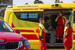 Kauempaa otettu ulkokuva pysäköitynä olevasta Etelä-Karjalan hyvinvointialueen ambulanssista, jossa sivuovi auki ja sisällä näkyy yksi puna-keltaisessa työasussa oleva ensihoitaja seisomassa ja puhumassa puhelimeen ja oviaukon sivuilla kaksi samanlaisiin asuihin pukeutunutta ensihoitajaa seisoo sivuittain.