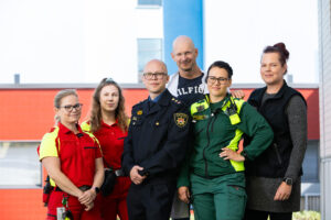 Ryhmäkuva kuudesta Etelä-Karjalan hyvinvointialueen työntekijästä eri ammateissa. Etualalla ensihoitajia punaisissa ja vihreissä asuissa sekä virkapukuinen pelastusalan työntekijä. Taustalla kaksi siviilivaatteissa olevaa henkilöä. Kuva on otettu ulkona sairaala rakennuksen edustalla