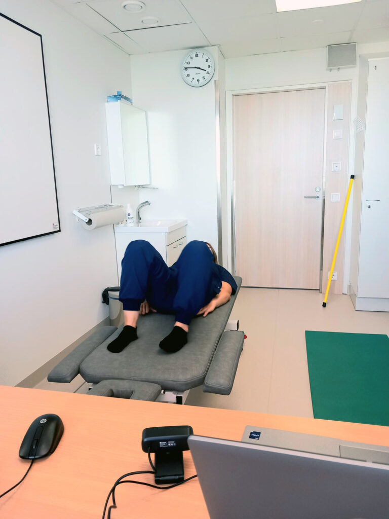 Tummansiniseen hoitajan asuun pukeutunut fysioterapeutti on selinmakuulla hoitosängyssä jalat koukussa. Hän on huoneessa, jossa kuvan etualalla on kannettava tietokone ja erillinen kamera, vasemmalla hoitosänky, taustalla on ovi huoneeseen. Huone on valoisa.