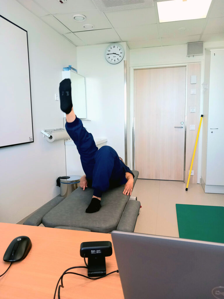 Tummansiniseen hoitajan asuun pukeutunut fysioterapeutti on selinmakuulla hoitosängyssä jalat koukussa. Toisen jalan hän on ojentanut suoraksi ylös. Hän on huoneessa, jossa kuvan etualalla on kannettava tietokone ja erillinen kamera, vasemmalla hoitosänky, taustalla on ovi huoneeseen. Huone on valoisa.
