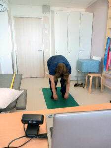 Tummahiuksinen ja lyhythihaiseen hoitajan asuun pukeutunut fysioterapeutti kumartaa syvään eteen selkä pyöreänä kädet maata kohti roikkuen. Hän on huoneessa, jossa kuvan etualalla on kannettava tietokone ja erillinen kamera, vasemmalla hoitosänky, taustalla ovi huoneeseen ja oikealla välineistöä. Huone on valoisa.