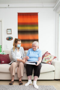 Nainen istuu sohvalla ikäihmisen kanssa ja katsoo papereita, joita on kansion päällä sylissä. Takana on värikäs ryijy seinällä.