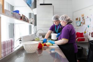 Kaksi henkilöä valmistamassa salaattia lilat paidat ja keittiömyssyt päässä