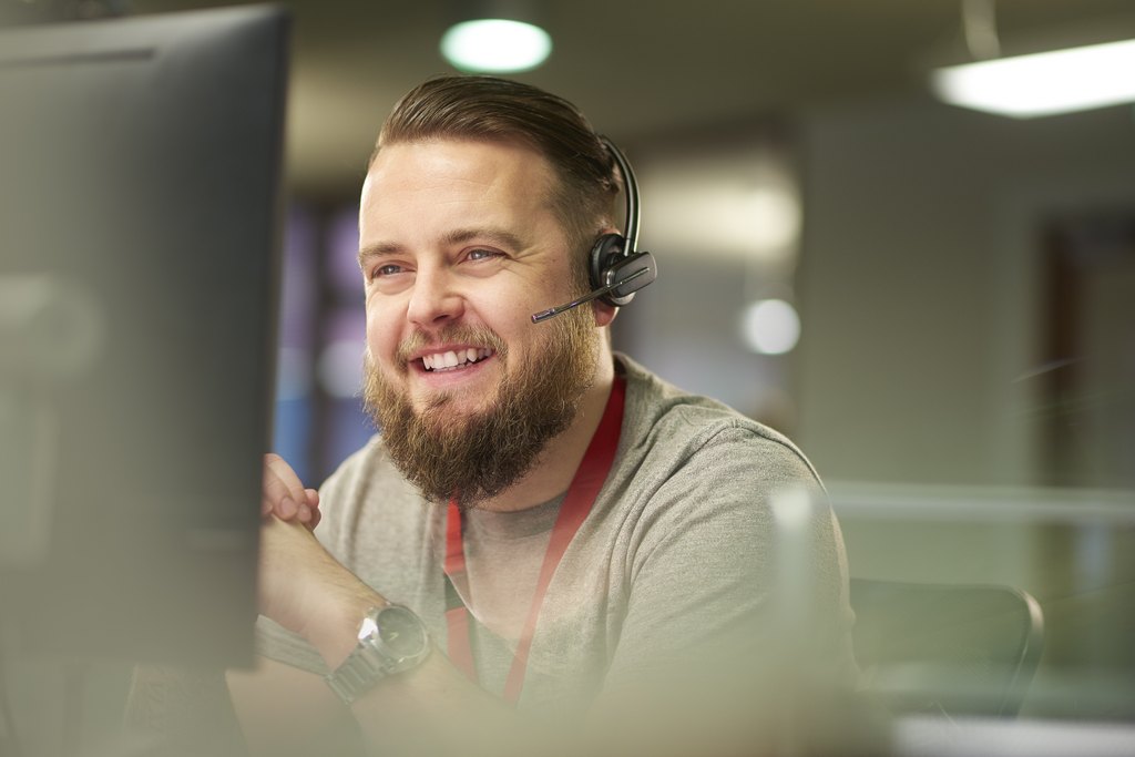 Mies istuu luurit päässä tietokoneen edessä ja hymyilee luurit päässä