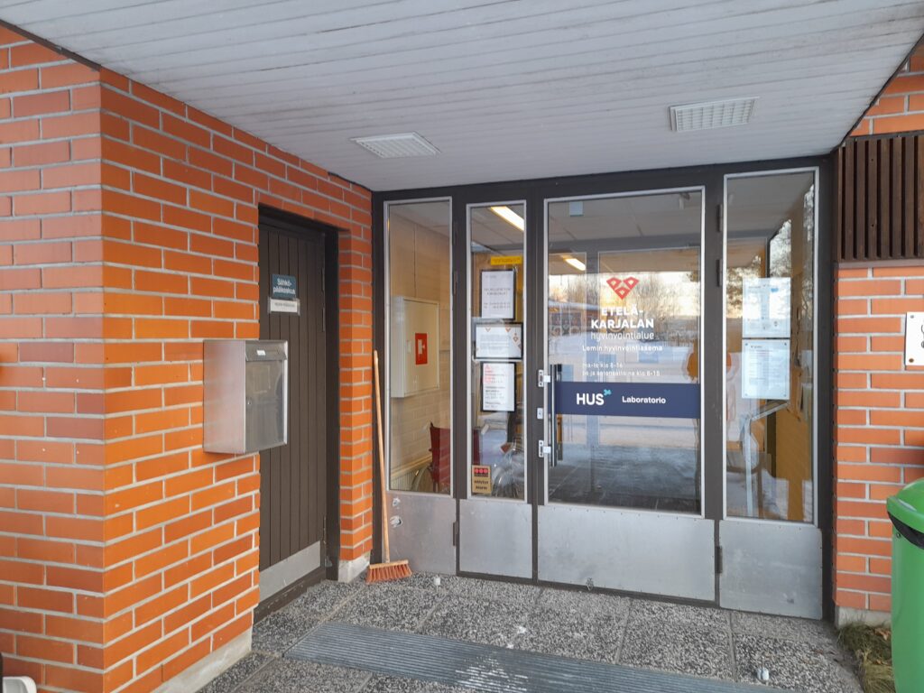 Tiilirakennuksen sisäänkäynti, jossa on lasiovet. Ovien lasipinnassa on tekstiä, jossa mainitaan 'Etelä-Karjalan hyvinvointialue', 'Lemin hyvinvointiasema' ja 'HUS Laboratorio'. Oikealla puolella on vihreä roskakori. Vasemmalla seinällä on metallinen postilaatikko ja ovi, jossa on kyltti 'Sähköpääkeskus'. Sisäänkäynnin edustalla on harmaa kuramatto, ja oven vieressä lattialla on punainen harja.