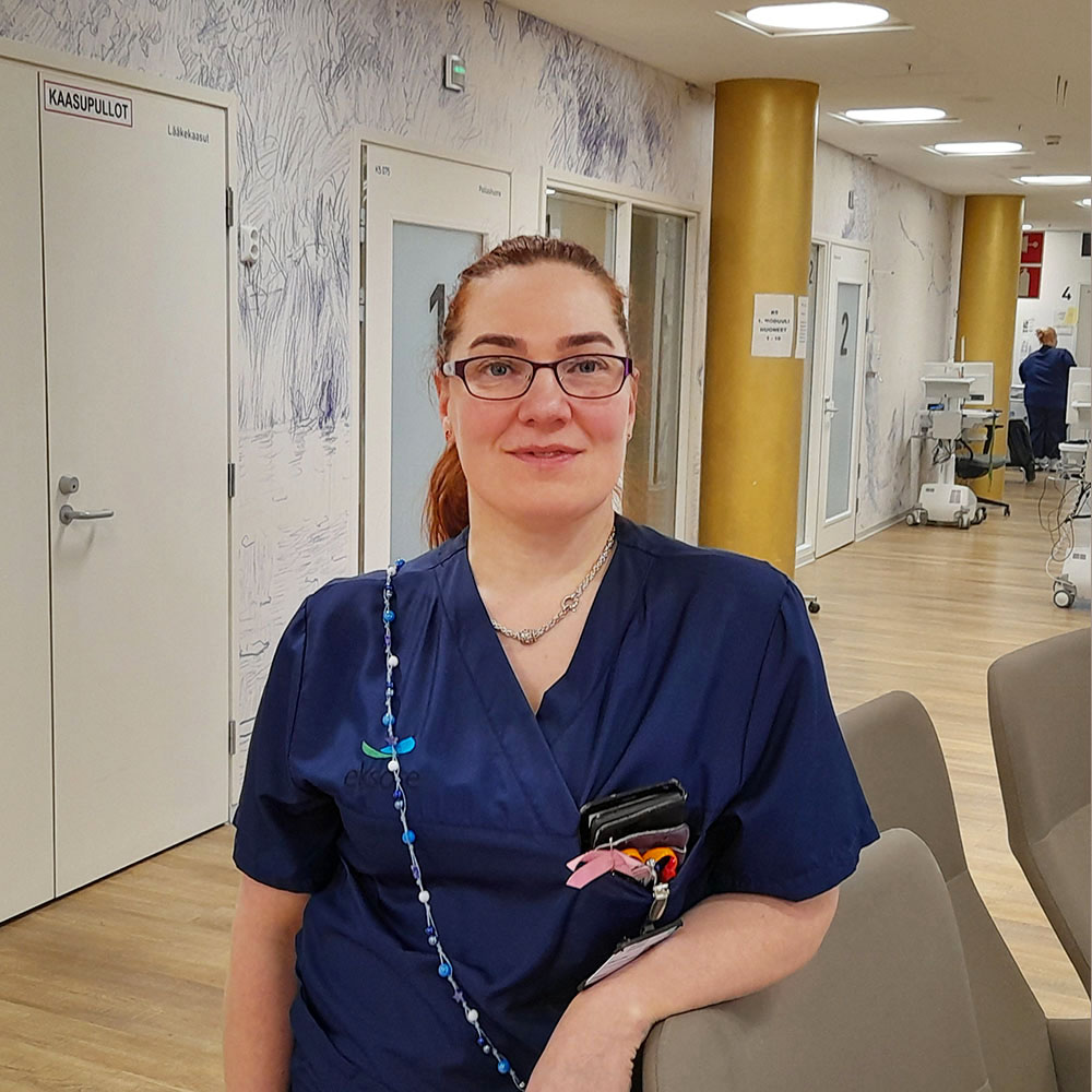 Nainen sairaanhoitajan tummansinisessä työasussa seisoo sairaalan vastaanottoalueella nojaten käsinojaan. Hänellä on ruskeat hiukset, silmälasit ja hymyilevä ilme. Taskussaan hänellä on työvälineitä ja kaulassaan helminauha. Taustalla näkyy sairaalaympäristö valkoisine ovineen ja hoitolaitteineen.