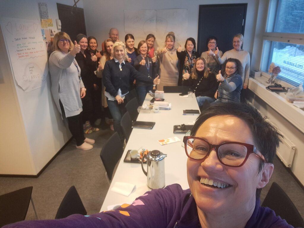 Ryhmä ihmisiä kokoushuoneessa hymyilemässä ja näyttämässä peukaloa ylös. Etualalla Nina Holopainen ottaa ryhmäselfien. Seinillä näkyy fläppitauluja, ja pöydällä on kahvikuppeja ja muistiinpanovälineitä.