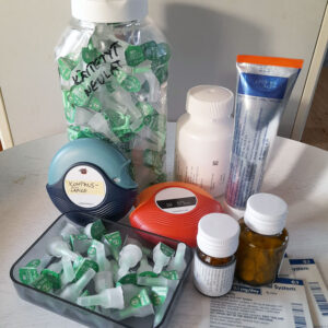 Pöydällä on erilaisia lääkinnällisiä tarvikkeita ja lääkkeitä. Kuvassa näkyy suuri muovipurkki, jossa on vihreäkorkkisia neuloja ja jossa on käsinkirjoitettu etiketti 'käytetyt neulat'. Sen edessä on kaksi pyöreää inhalaattoria, joista toisessa on etiketti 'kohtauslääke'. Lisäksi pöydällä on useita lääkepulloja, voideputkilo, lääkelaastareita ja avoin rasia, jossa on lisää neuloja ja muita tarvikkeita.