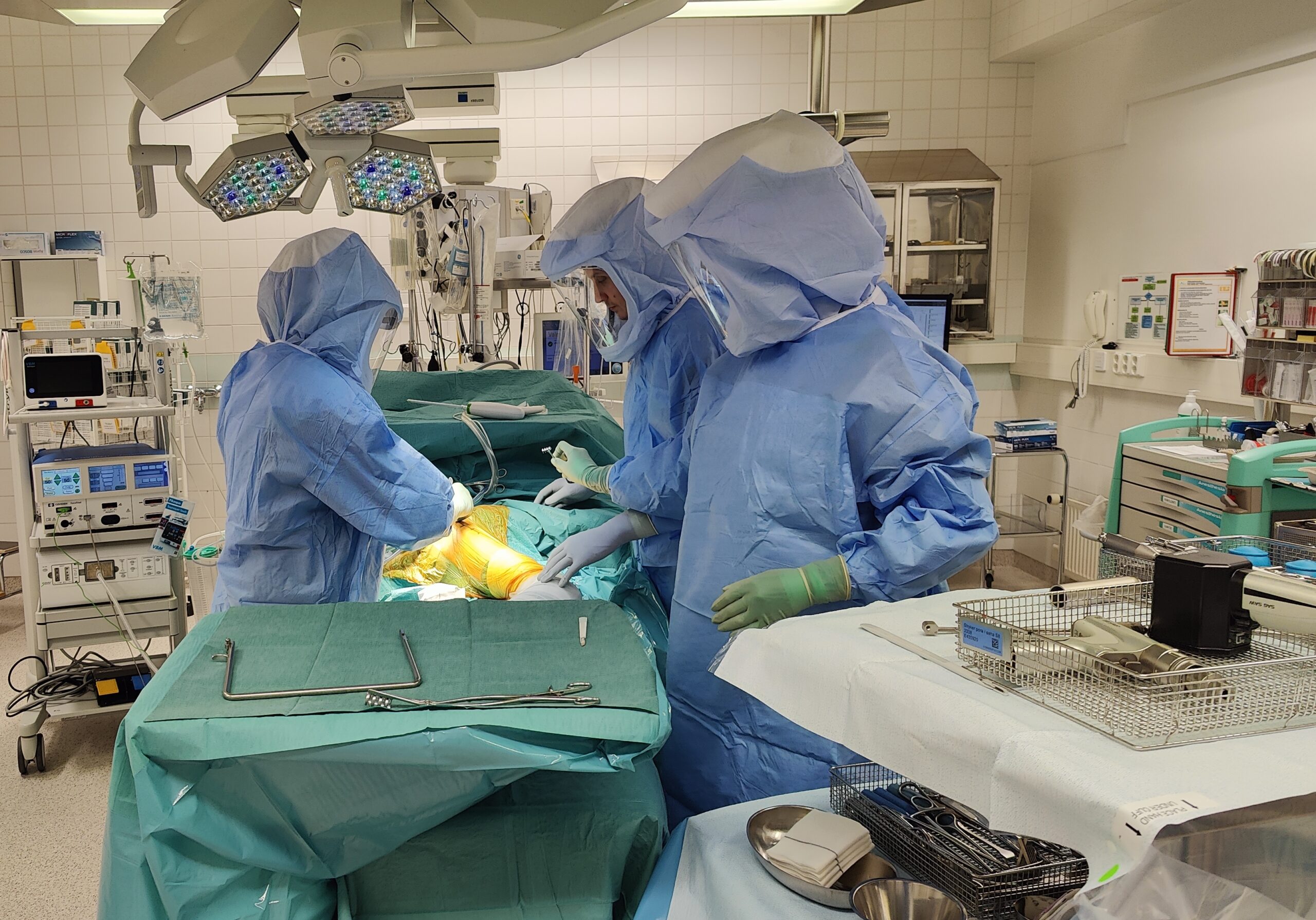 Leikkaussalissa kaksi kirurgia ja avustava henkilö työskentelevät steriilisti pukeutuneina sinisissä suojavarusteissa ja kokovisiireissä. Potilaan jalka on esillä ja suojattu steriilillä keltaisella kalvolla. Leikkaussalissa on modernia lääketieteellistä laitteistoa, kirurgisia instrumentteja ja vihreä leikkausliina peittämässä potilaan muuta kehoa.