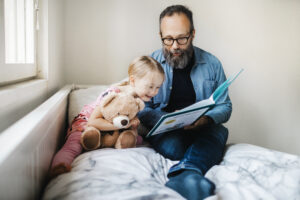 Isä lukee lapselleen kirjaa sängyssä. Tyttölapsella on nalle sylissään.