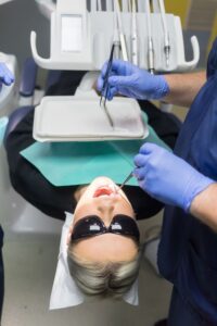 Potilas makaa hammashoitotuolissa aurinkolasit silmillään hammaslääkärin tehdessä suun tarkastusta. Hammaslääkäri ja avustaja käyttävät sinisiä suojakäsineitä ja työskentelevät hammashoitolaitteiden äärellä.