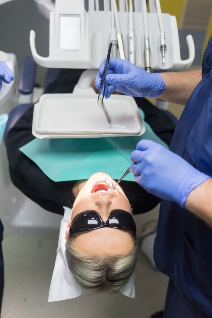 Potilas makaa hammashoitotuolissa aurinkolasit silmillään hammaslääkärin tehdessä suun tarkastusta. Hammaslääkäri ja avustaja käyttävät sinisiä suojakäsineitä ja työskentelevät hammashoitolaitteiden äärellä.