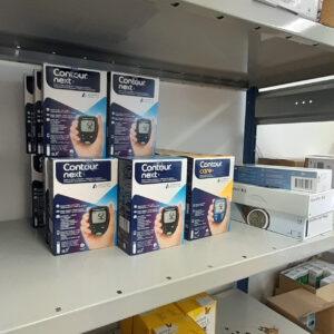 Hylly, jolla on pinottuna useita "Contour next" ja "Contour care" -verensokerimittareiden pakkauksia. Vieressä näkyy muita lääkinnällisiä tarvikelaatikoita ja alemmalla hyllyllä lisää tuotteita.