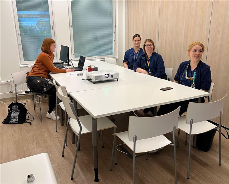 Neljä naista istuu valkoisen neuvottelupöydän ääressä modernissa kokoushuoneessa. Yksi heistä, ruskeaan puseroon pukeutunut nainen, käyttää kannettavaa tietokonetta. Kolme muuta naista, jotka näyttävät olevan terveydenhuollon ammattilaisia, istuvat pöydän toisella puolella, pukeutuneina tummansinisiin työasuihin. Pöydällä on projektori, kynäteline ja muita toimistotarvikkeita. Taustalla on ikkunoita, joissa on kaihtimet, sekä puuseinäpaneeli.