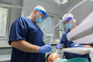 Kaksi suun terveydenhuollon ammattilaista hoitaa potilasta hammaslääkärin vastaanotolla. Molemmilla on päällään siniset työasut, kasvomaskit ja kasvovisiirit. He käyttävät suunhoitovälineitä potilaan suussa, ja potilas makaa hoitotuolissa silmälaput silmillään.