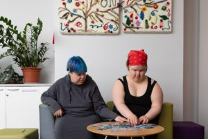 Kaksi nuorta naista istuu vierekkäin matalan pyöreän pöydän ääressä ja kokoaa palapeliä. Vasemmalla oleva henkilö käyttää harmaata hupparia ja hänellä on lyhyet sinimustat hiukset. Oikealla oleva henkilö pukeutuu mustaan hihattomaan paitaan ja punaiseen huiviin, joka on sidottu päähän. Molemmat keskittyvät palapelin kokoamiseen. Taustalla näkyy valkoinen seinä, jolla on kaksi tekstiilitaideteosta, joissa on puun oksia ja värikkäitä koristeita. Vasemmalla puolella huoneessa on vihreä kasvi ruukussa ja valkoinen kaappi.