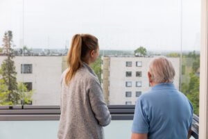 Nainen seisoo ikäihmisen kanssa parvekkeella ja katsoo maisemia selät ovat kameraan päin