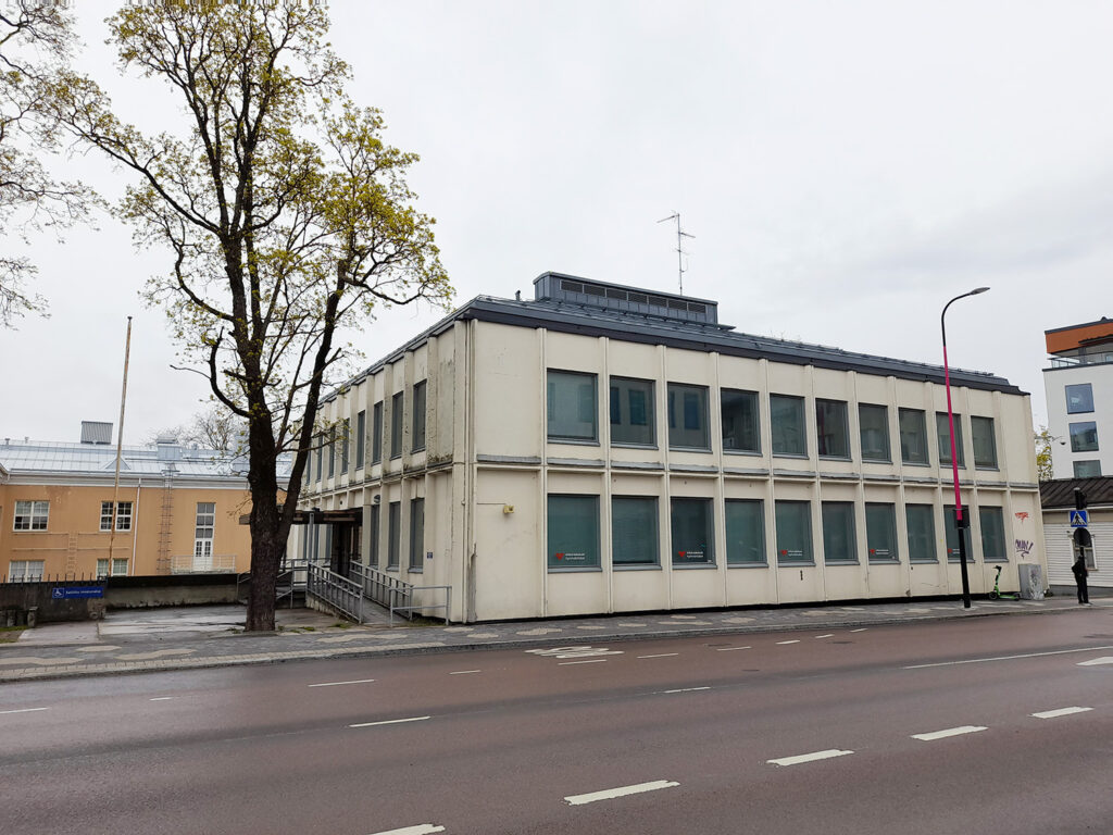Vaalea kaksikerroksinen virastomainen rakennus kadun varrella harmaana ja sateisena päivänä. Rakennuksessa on useita samanlaisia ikkunoita, joissa on sälekaihtimet. Etualalla näkyy jalkakäytävä ja ajotie, jossa on valkoisia kaistaviivoja. Rakennuksen edessä on esteetön luiska, ja vasemmalla kasvaa iso lehtipuu. Taustalla näkyy lisää rakennuksia, muun muassa vaaleankeltainen ja moderni lasipintainen talo.