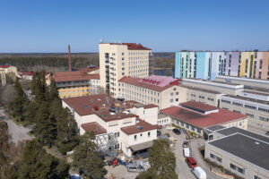 Ilmakuva sairaala-alueesta aurinkoisena päivänä. Etualalla näkyy vaaleita sairaalarakennuksia punaisine kattoineen, joiden ympärillä on meneillään rakennustyömaa. Taustalla kohoaa korkea kerrostalo sekä useita muita sairaalakompleksin rakennuksia. Oikeassa reunassa näkyy värikäs, monivärinen rakennus. Taustalla metsämaisemaa ja vesistö.