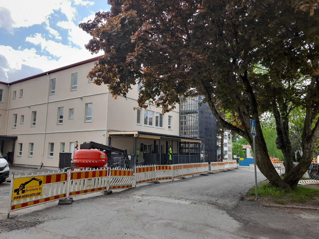 Sairaalan ulkopuolinen näkymä, jossa rakennuksen edessä on rakennustyömaa-alue. Etualalla on valkoinen rakennus, jonka edessä on työmaa-aita ja punainen työmaakone. Vasemmalla näkyy sisäänkäynti, ja rakennuksen oikealla puolella on iso puu ja suojatie.