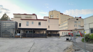 Panoraamakuva vaaleasta rakennuksesta, jonka edessä on harmaa hiekkainen piha-alue. Rakennuksessa on tiloja eri tasoissa. Rakennuksen edessä on tummanharmaat