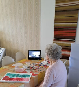 Ikäihminen istuu pöydän ääressä ja katsoo tablettia, edessään pöydällä on maalaus ja maalaustarvikkeita