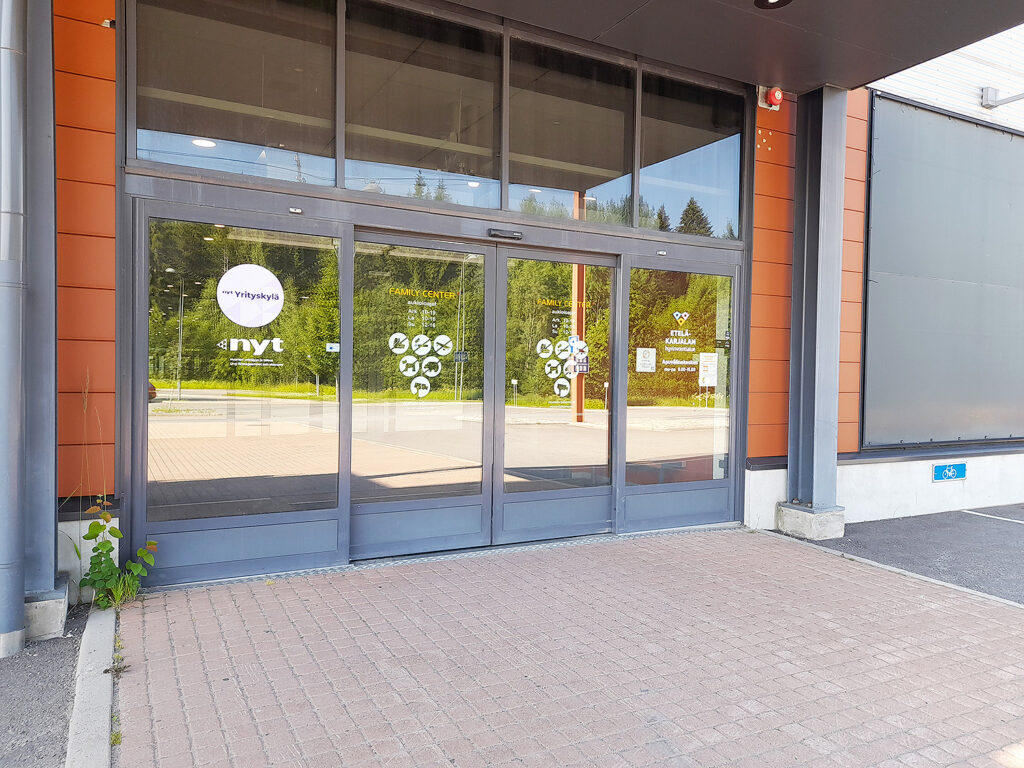 Sisäänkäynti Kauppakeskus Family Centeriin. Kuvassa lasiset liukuovet, joissa valkoisia tarratekstejä, kuten Etelä-Karjalan hyvinvointialue ja Yrityskylä. 