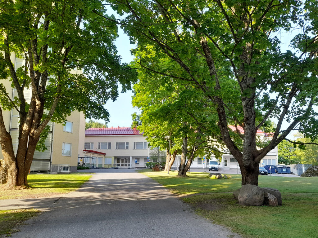 Puutarhamainen pihatie, jota reunustavat suuret vihreät puut. Taustalla näkyy vaalea rakennus punaisella katolla, ja oikealla on kiviryhmä nurmikolla.