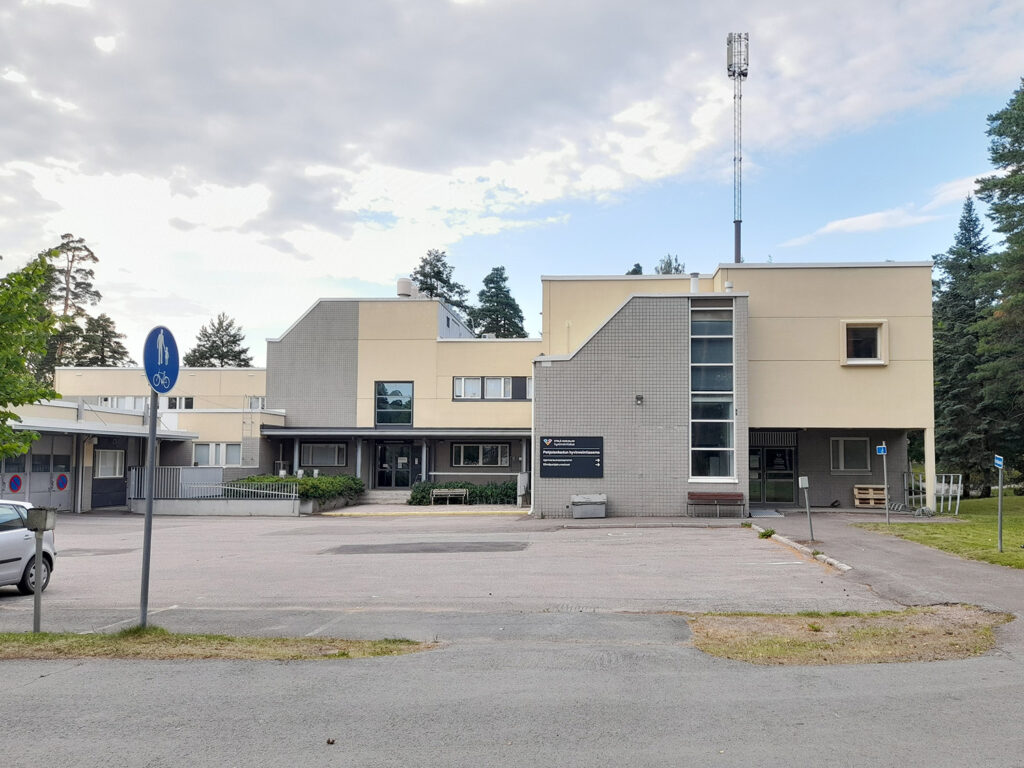 Vaalea julkinen rakennus, jonka edustalla on parkkialue. Rakennuksen edessä on kyltti, jossa lukee "Pohjolankadun hyvinvointiasema" ja muuta tekstiä. Taustalla metsää ja telemastorakennelma