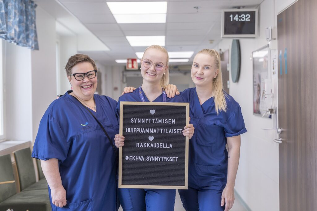 Kolme hymyilevää sairaanhoitajaa tai kätilöä seisoo sairaalan käytävällä sinisissä työasuissa. Keskimmäinen henkilö pitelee kylttiä, jossa lukee: "❤️ Synnyttämisen huippuammattilaiset ❤️ Rakkaudella @ekhva.synnytykset". Taustalla näkyy sairaalan käytävä, huoneiden ovia ja digitaalinen kellonäyttö, jossa lukee 14:32.