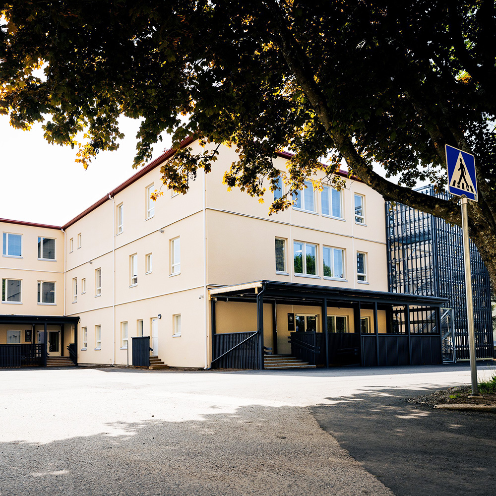 Vaaleankeltainen, kolmikerroksinen rakennus, jossa on useita ikkunoita ja kaksi sisäänkäyntiä katetulla rampilla. Etualalla on asfalttipiha, ja rakennusta varjostaa suuri lehtipuu. Oikealla näkyy suojatien liikennemerkki.