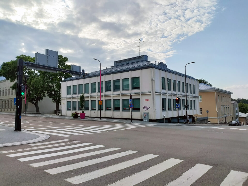 Keskustan risteys, jossa on useita suojateitä ja liikennevalot. Kuvassa näkyy valkoinen toimistorakennus, jonka julkisivussa on sälekaihtimet ja hieman graffiteja. Taivas on pilvinen.