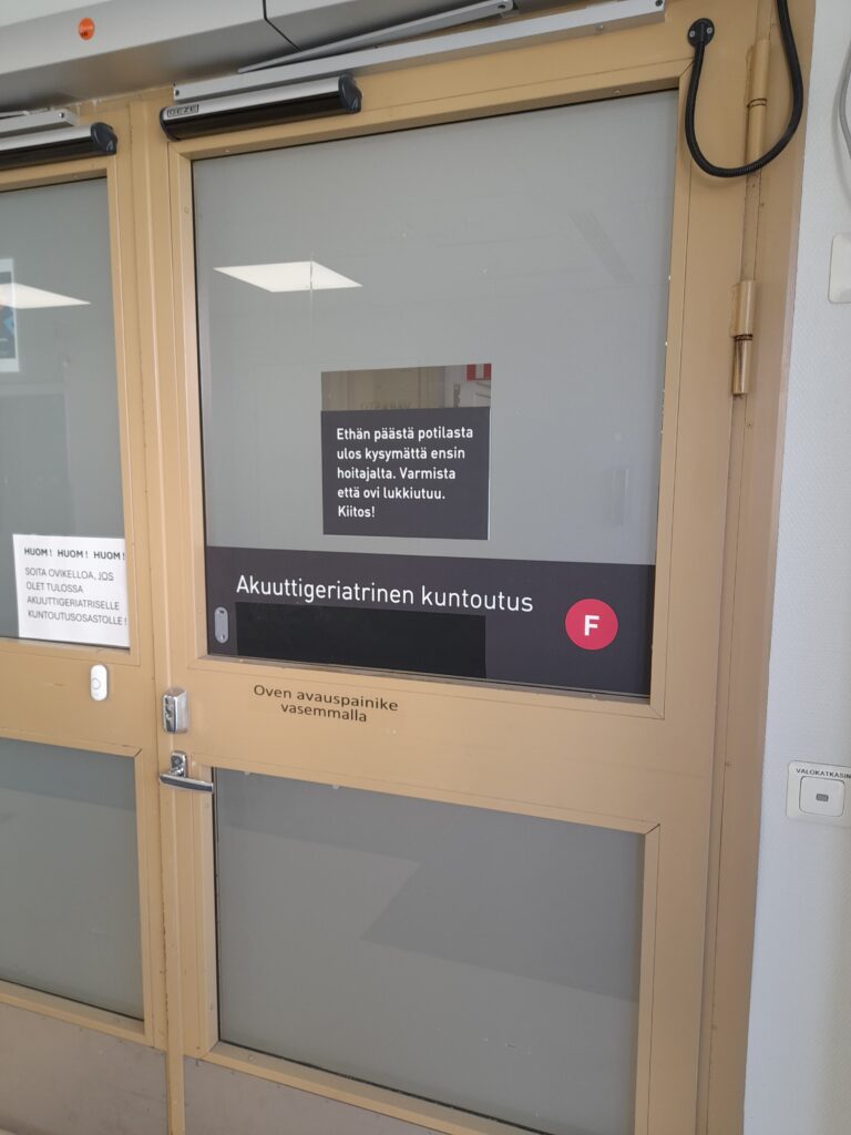 Sairaalan suljettu lasiovi, jossa on kyltti: 'Ethän päästä potilasta ulos kysymättä ensin hoitajalta. Varmista että ovi lukkiutuu. Kiitos!' Oven yläosassa lukee 'Akuuttigeriatrinen kuntoutus'. Oven vasemmalla puolella on painike oven avaamiseksi.