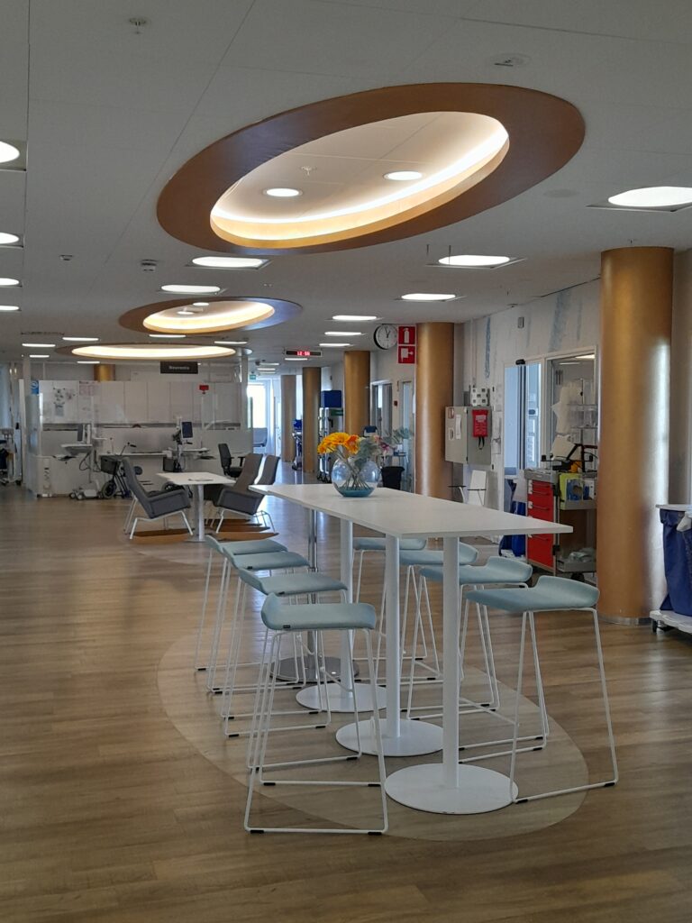 Sairaalan valoisa yhteistila, jossa on korkea valkoinen pöytä ja useita vaaleansinisiä baarijakkaroita. Taustalla näkyy nojatuoleja, hoitovälineitä ja huoneiden ovia. Katossa on suuria pyöreitä valoelementtejä ja pylväitä, jotka korostavat modernia sisustusta.