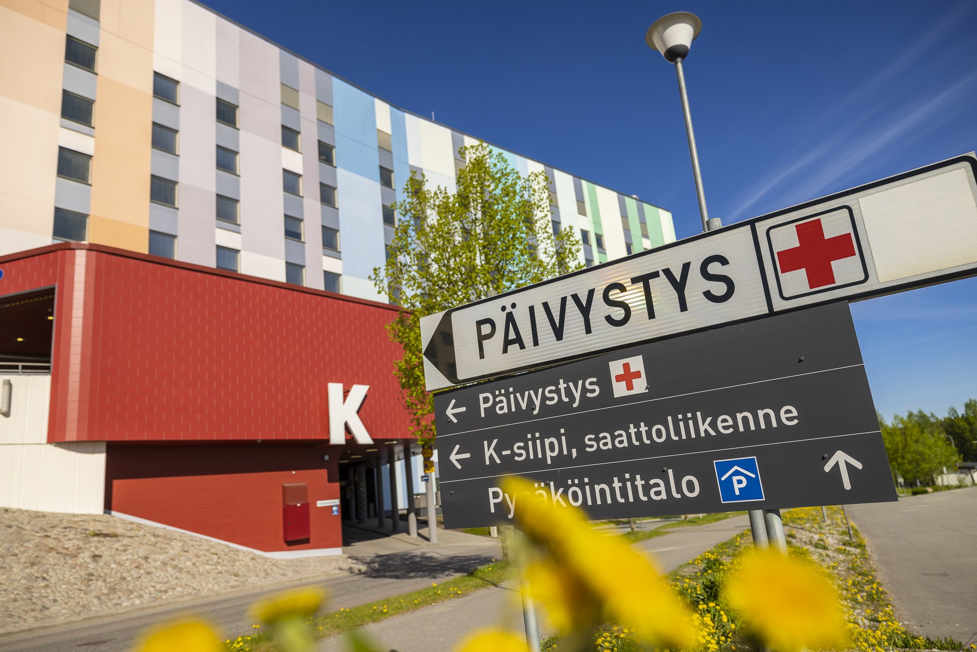 Sairaalan edustalla oleva kyltti, jossa lukee 'Päivystys' punaisen ristin symbolin kanssa. Taustalla näkyy monivärinen sairaalarakennus, punainen K-ovi ja sininen taivas. Etualalla on keltaisia kukkia.