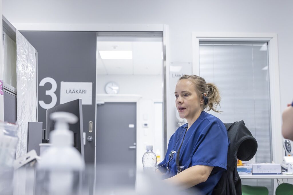 Sairaalan vastaanotossa työskentelevä hoitaja tai lääkäri istuu työpisteellä kuulokemikrofoni päässä ja puhuu. Taustalla näkyy ovi, jossa lukee 'Lääkäri', sekä toimiston kalusteita ja välineitä.