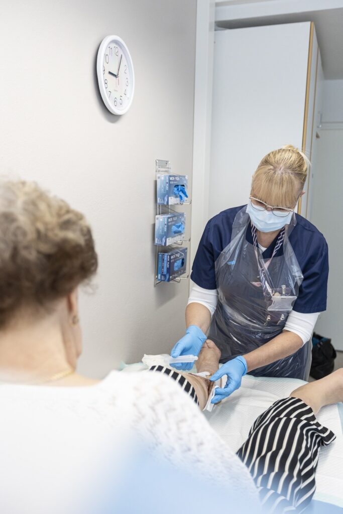 Hoitaja pukeutuneena siniseen työasuun, muoviseen suojaliinaan, suojamaskiin ja suojakäsineisiin hoitaa potilaan jalkaa sairaalaympäristössä. Potilas istuu selkä kameraan päin yllään valkoinen pitsipusero ja mustavalkoraidalliset housut. Taustalla seinällä näkyy kello ja hyllyssä kertakäyttökäsinepaketteja.