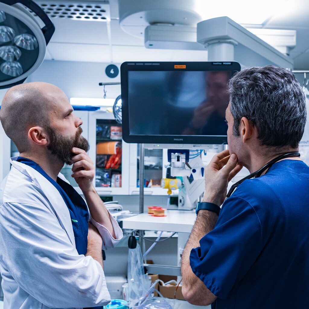 Kaksi lääkäriä seisoo sairaalaympäristössä tutkimassa ja pohtimassa tietokoneen näyttöä. Vasemmalla oleva lääkäri on kalju ja partasuinen, pukeutunut valkoiseen takkiin ja siniseen työasuun. Oikealla oleva lääkäri on harmaantuvahiuksinen ja pukeutunut tummansiniseen työasuun, kaulassaan stetoskooppi. Molemmat pitävät kättään leuallaan miettivästi. Taustalla näkyy sairaalalaitteita ja tarvikkeita.