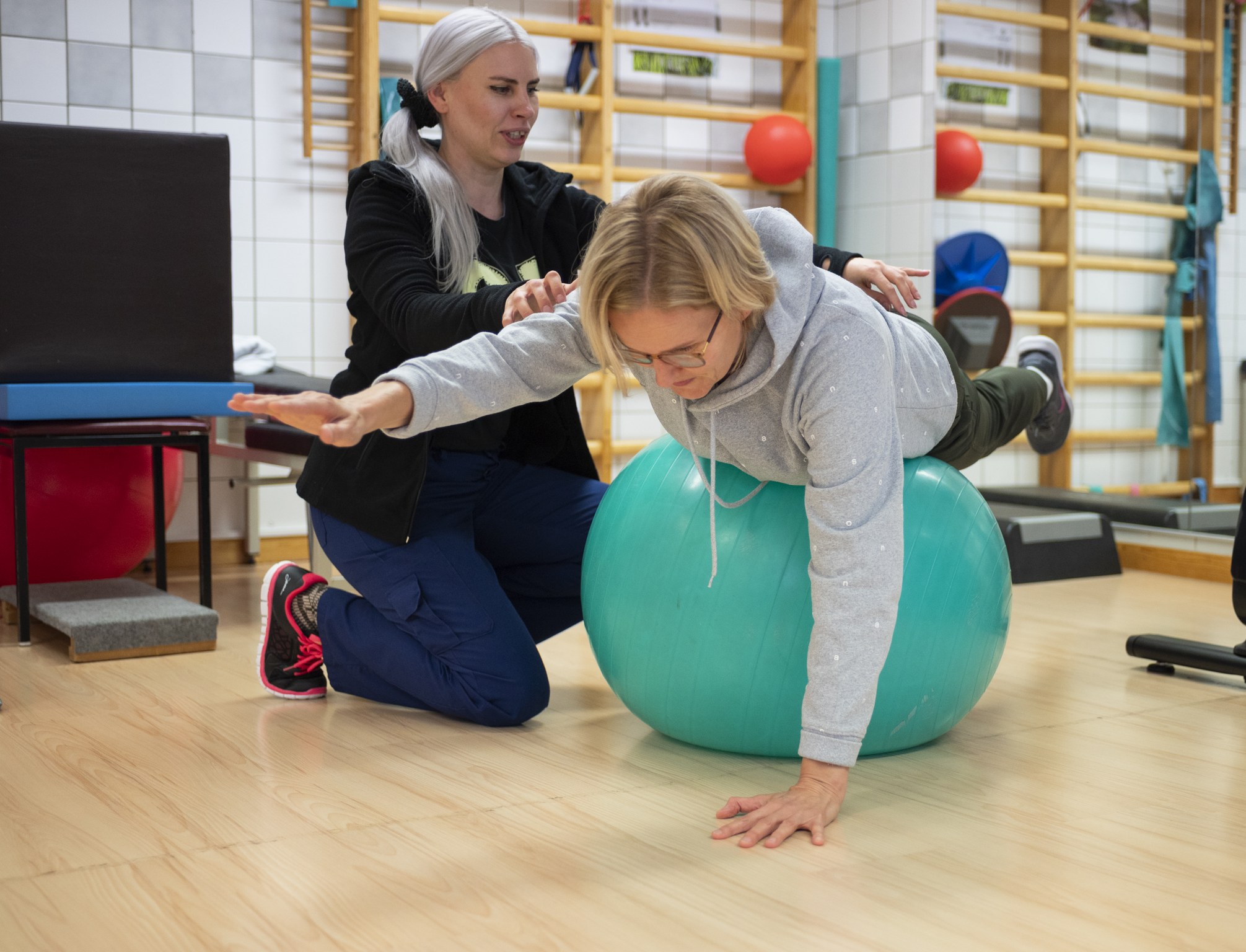 Fysioterapeutti ohjaa asiakasta harjoituksessa, jossa asiakas makaa vatsallaan jumppapallon päällä ja ojentaa käsiään eteenpäin tasapainon ylläpitämiseksi.