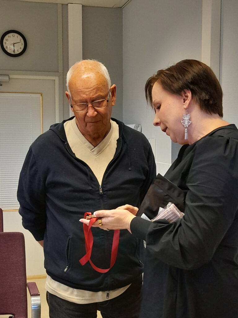 Vanhempi mies ja nainen seisovat sisätiloissa. Nainen ojentaa miehelle punaisella nauhalla varustetun mitalin, ja molemmat katsovat esinettä yhdessä.