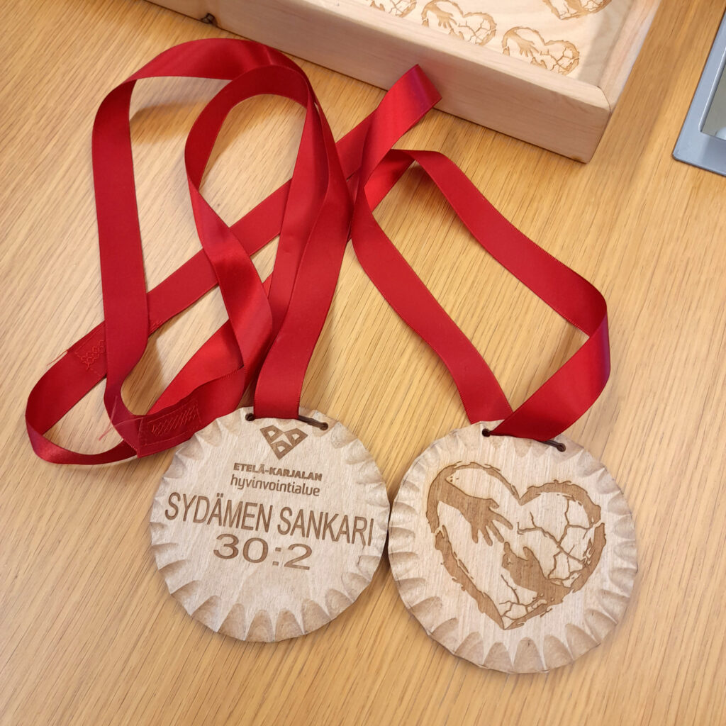 Pöydällä olevat puiset koristeelliset mitalit, joissa punainen kiiltävä kaulanauha. Mitalin toisella puolella on teksti Sydämen sankari 30:2 ja toisella puolella tyylitelty sydän ja toisiaan koskettavat kädet. 