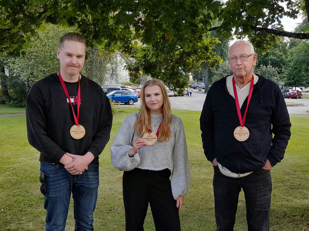 Kolme henkilöä seisoo ulkona nurmikolla puiden alla. Kaikilla on kaulassaan puiset mitalit punaisessa nauhassa.