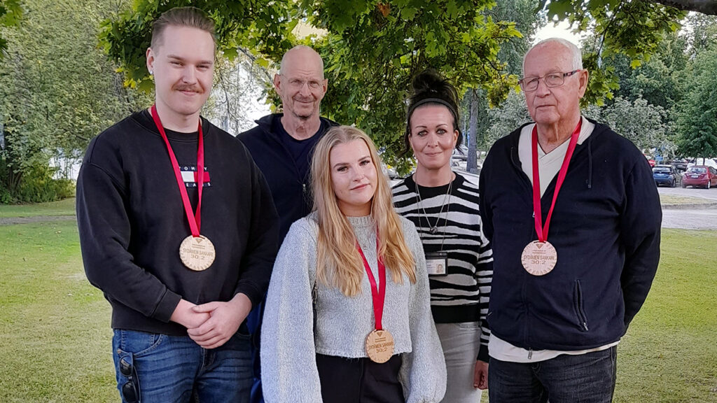Viisi henkilöä seisoo ulkona nurmikolla puiden alla. Kolmella heistä on kaulassaan puiset mitalit punaisessa nauhassa.