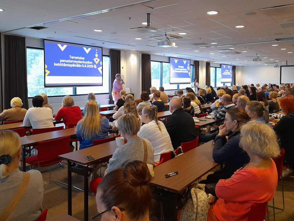 Takaapäin kuvattu suuri kokoustila, joka on aivan täysi istuvista ihmisistä. Edessä on nainen puhumassa mikkiin. Kolmelle isolle led-näytölle on heijastettu teksti Tervetuloa perusterveydenhuollon kehittämispäivään 5.9.2025.