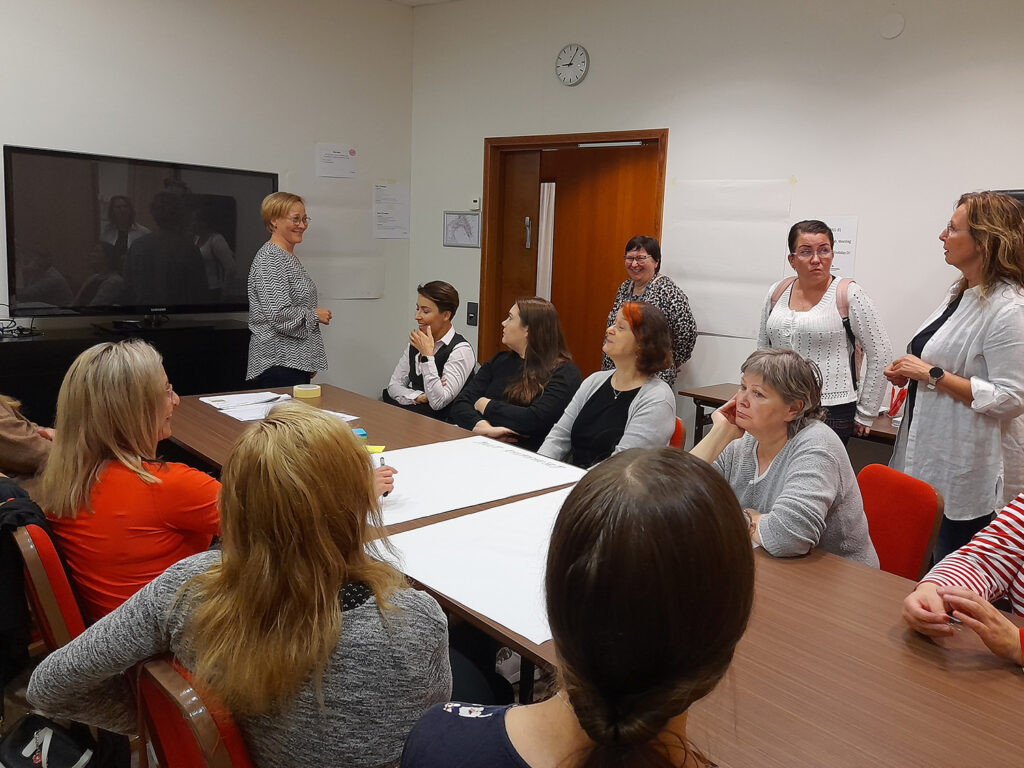 Pienemmässä kokoustilassa kymmenkunta naista istuu pitkän kokouspöydän äärellä. Ryhmän vetäjä ja kolme muuta naista seisovat. Kuvasta välittyy iloinen tilanne hymyjen perusteella.